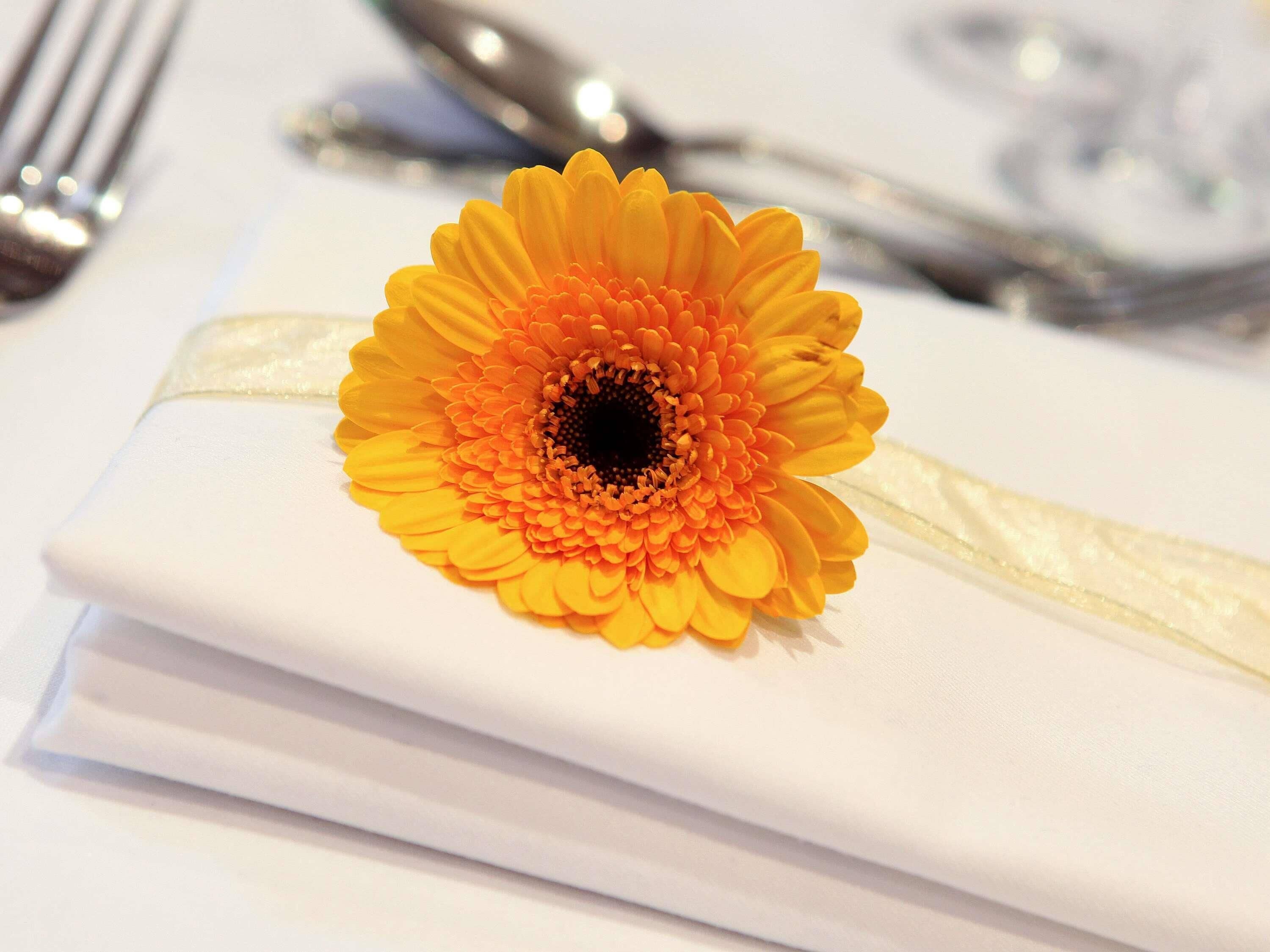 A table setting with a white tablecloth, a folded napkin, and a yellow flower boutonniere.