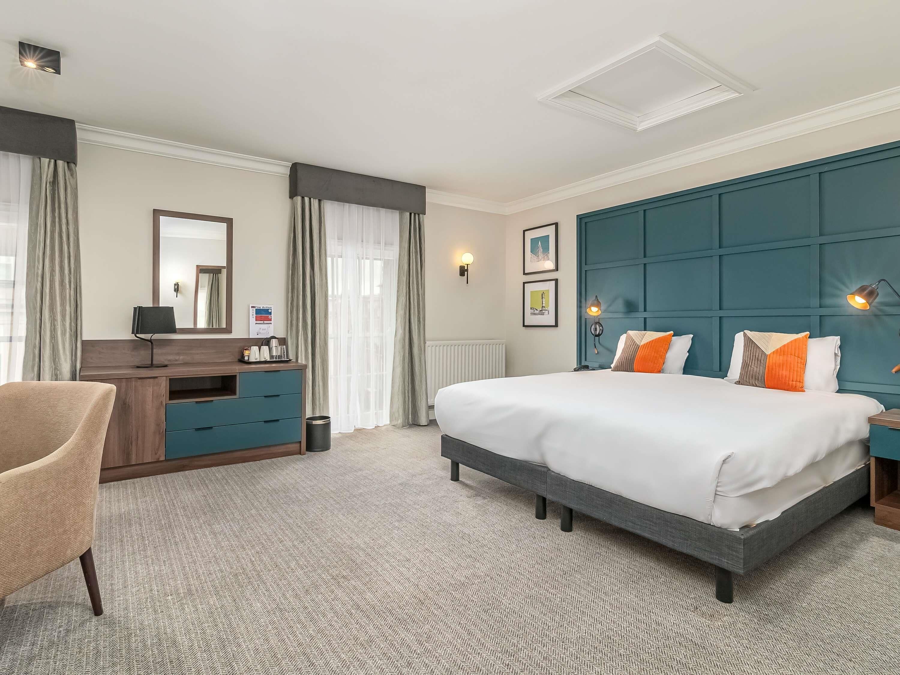 Una habitación de hotel con cama, escritorio, silla y un gran ventanal con cortinas.