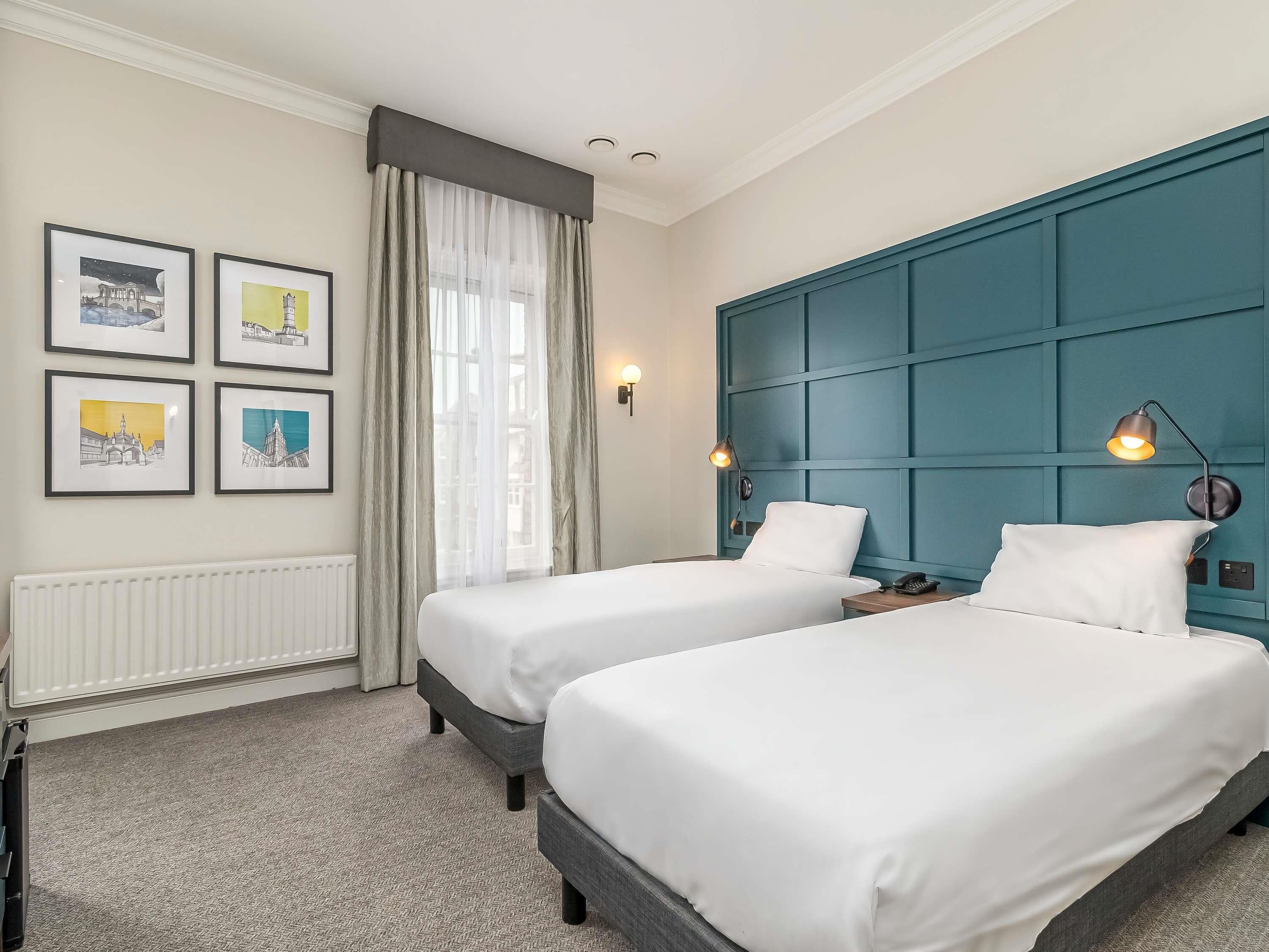 A hotel room with two beds, a radiator, a window with sheer curtains, and framed pictures on the wall.