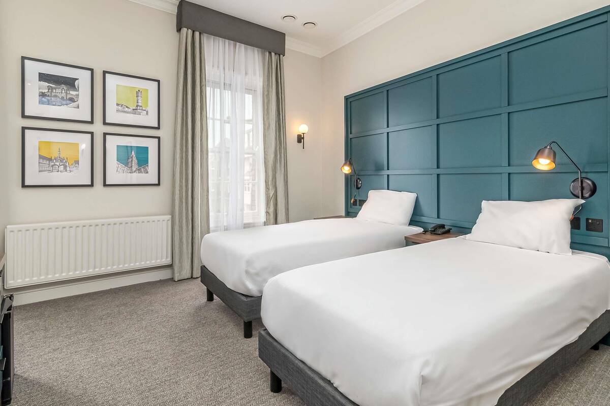 A hotel room with two beds, a radiator, a window with sheer curtains, and framed pictures on the wall.