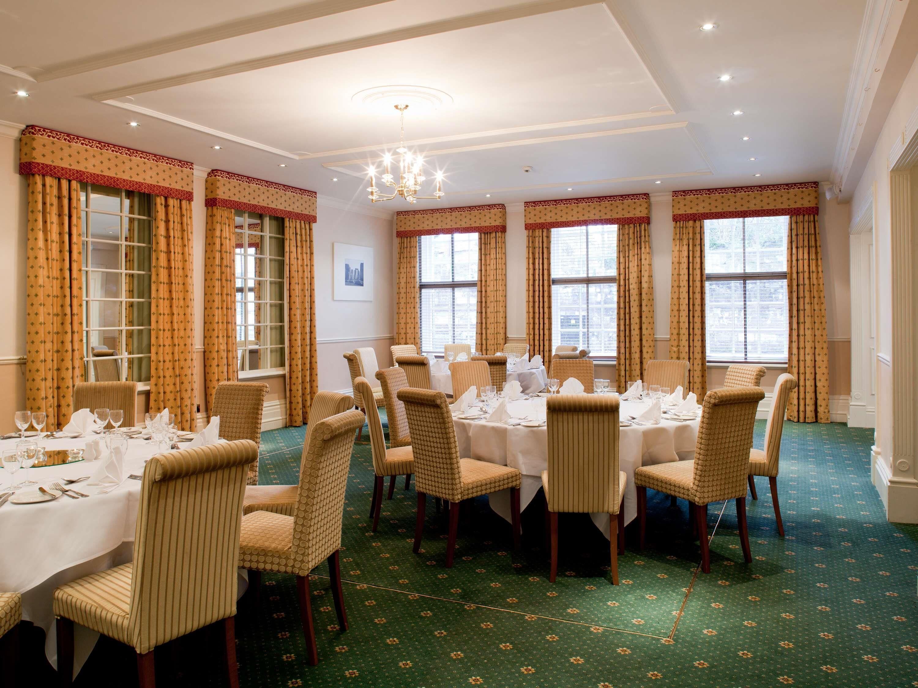 A formal dining room with multiple tables set for service, large windows with curtains, and a chandelier.