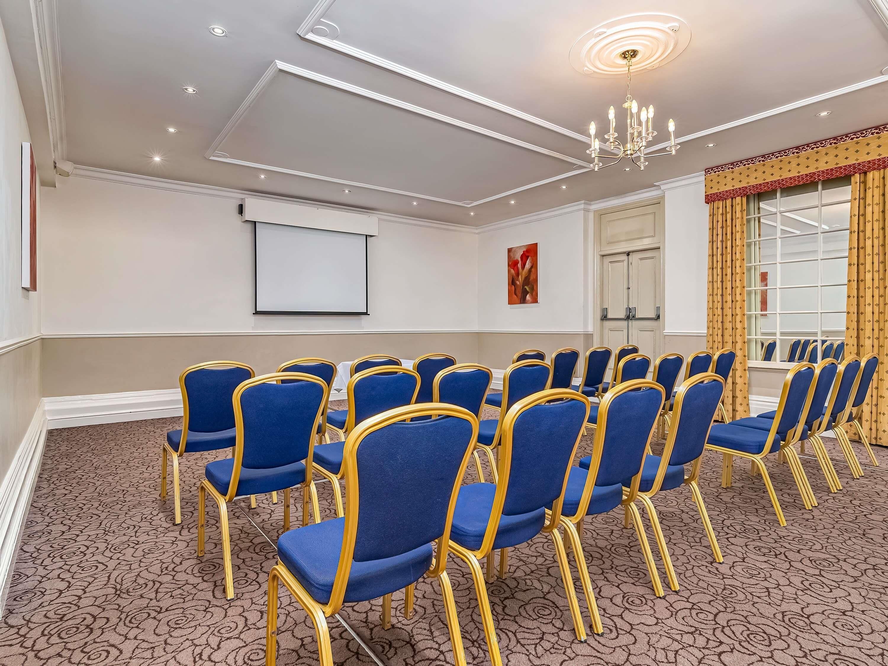 A conference room with rows of blue chairs, a projector screen, a chandelier, and patterned carpet.