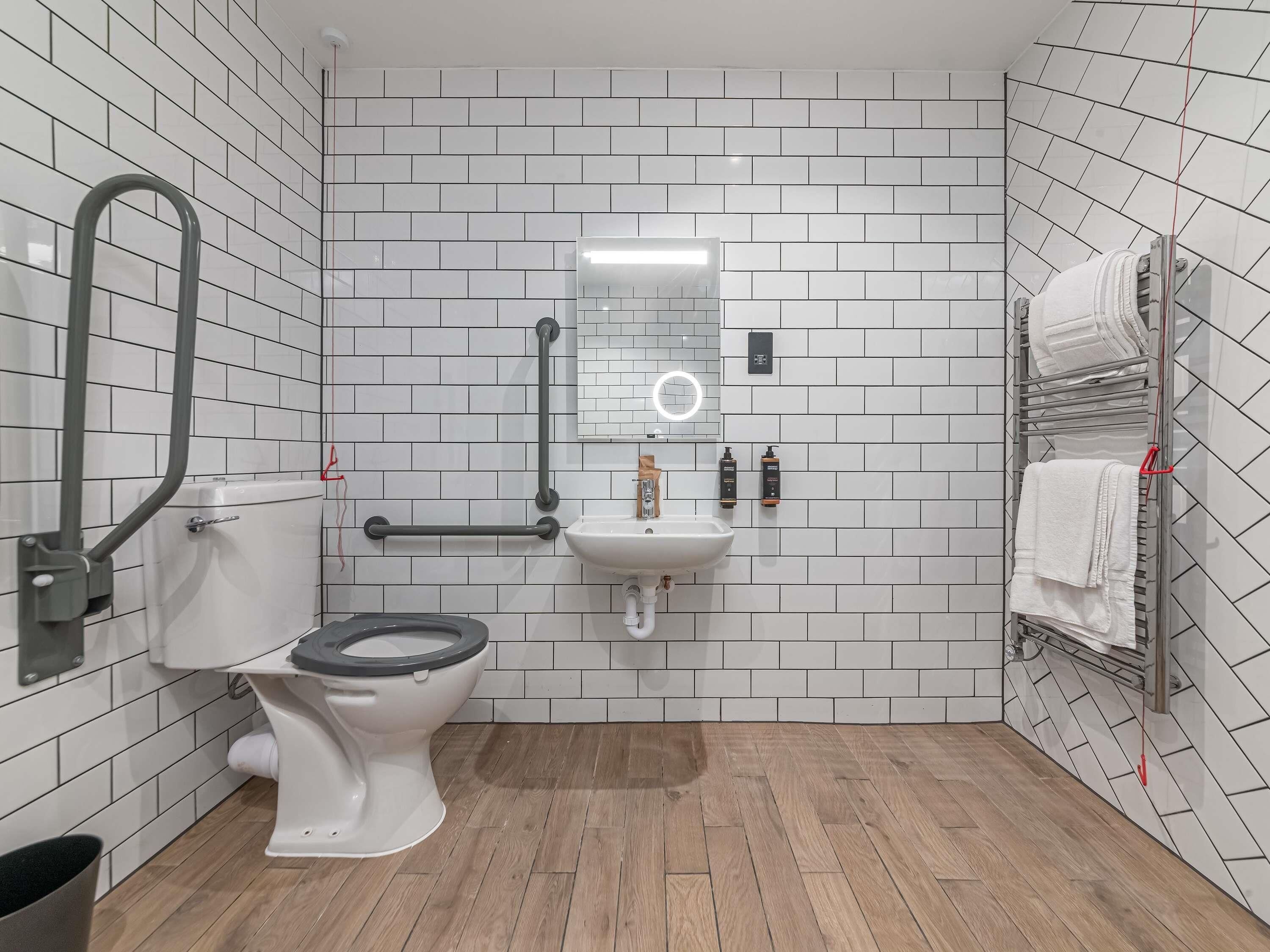 A modern bathroom with white tiled walls, a wooden floor, a toilet, a sink, a mirror, and a towel rack.