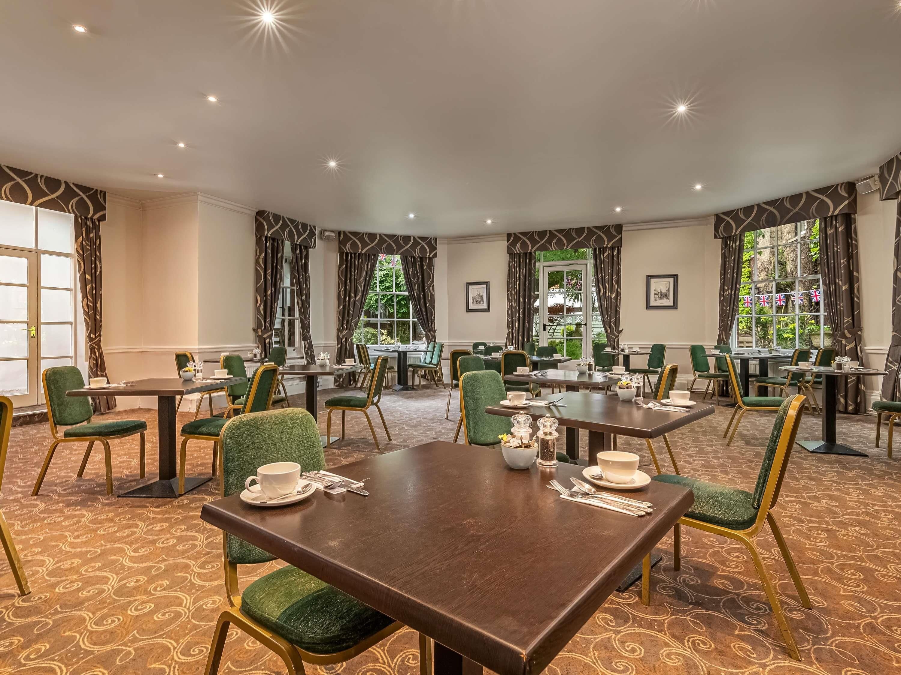 Une salle à manger d’hôtel avec de nombreuses tables et chaises, de grandes fenêtres avec des rideaux et un tapis à motifs.
