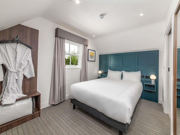 A hotel room with a large bed, a robe rack, a window with curtains, a headboard, and a nightstand with a lamp.