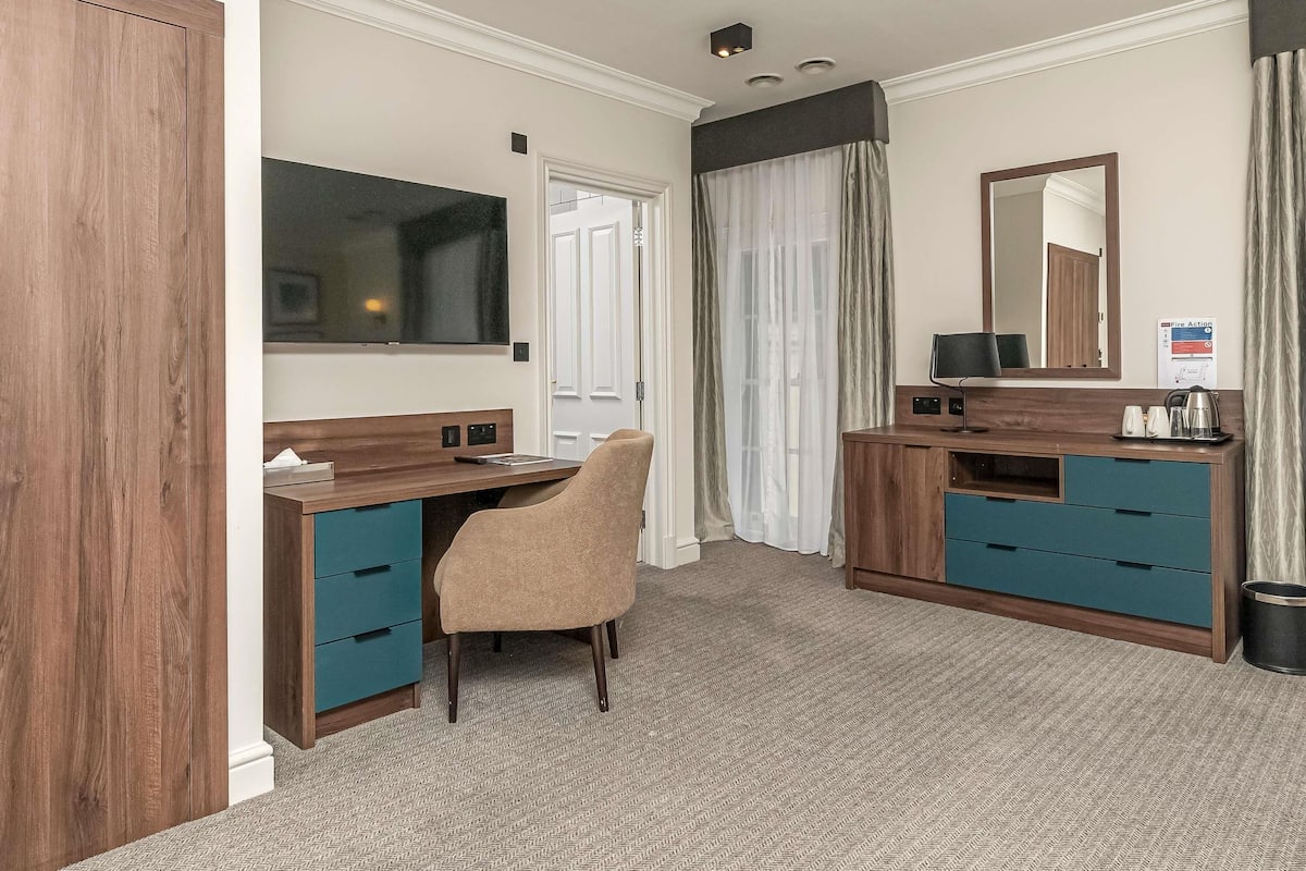A hotel room with a wooden desk, a chair, a TV, a mirror, and a 'Fire Action' sign.