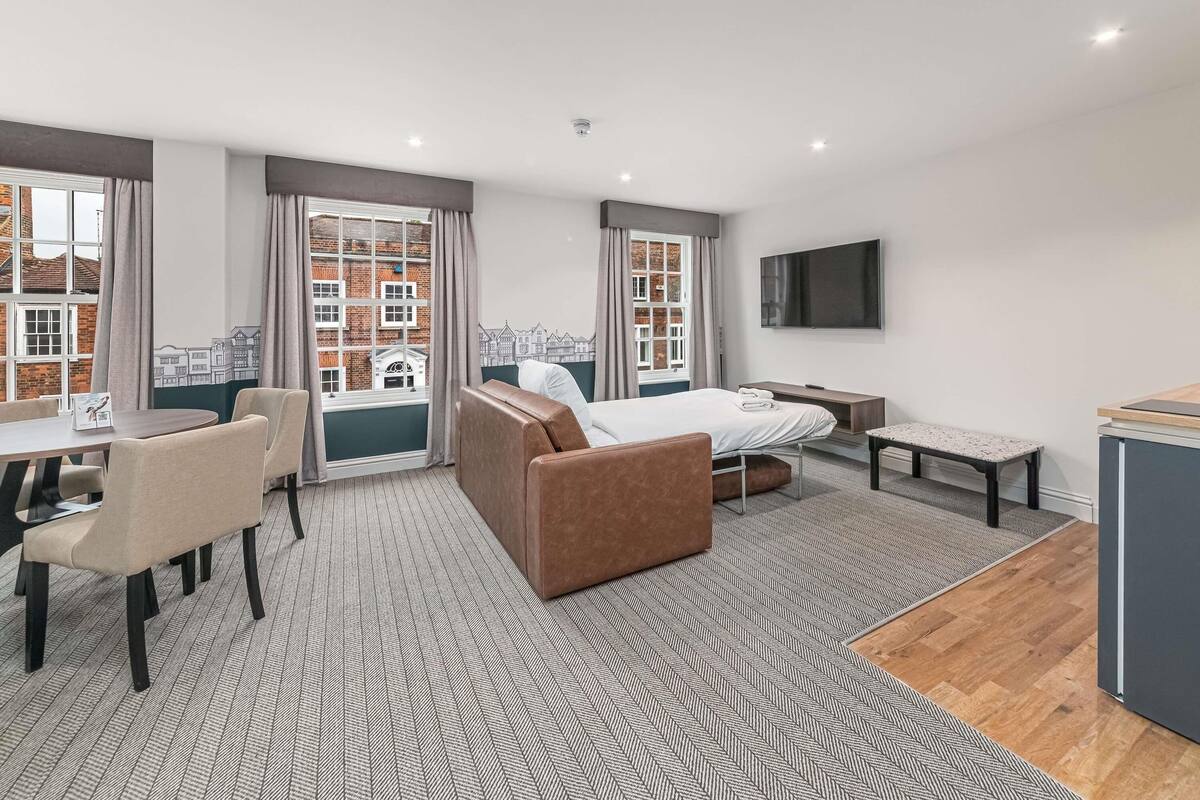 A hotel room with a bed, a desk, a chair, a TV, and a view of buildings through the window.