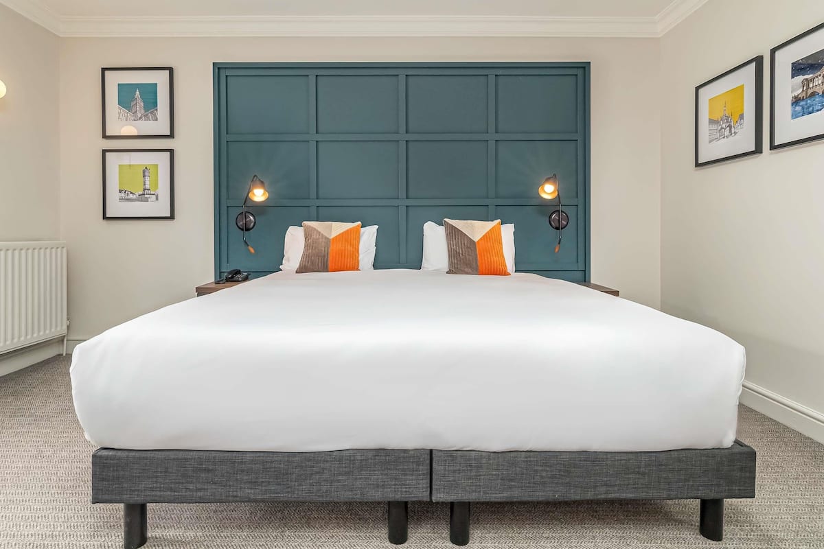 Habitación de hotel con una cama grande, dos lámparas de noche, cabecera y cuadros en las paredes.