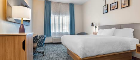 A hotel room with a bed, a desk, a chair, a lamp, a window with curtains, and framed pictures on the wall.