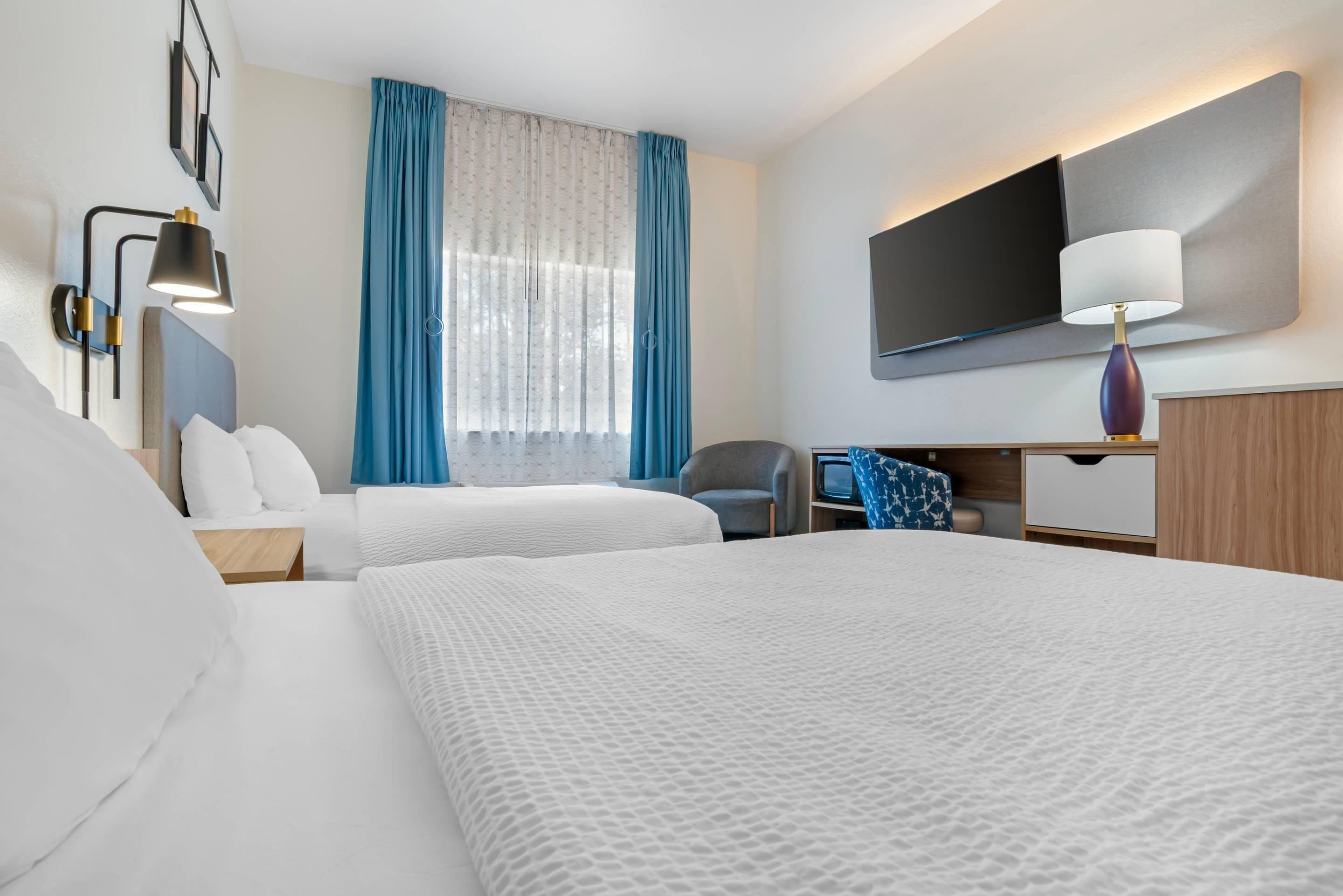 A hotel room with two beds, a desk, a chair, a TV, and a window with curtains.