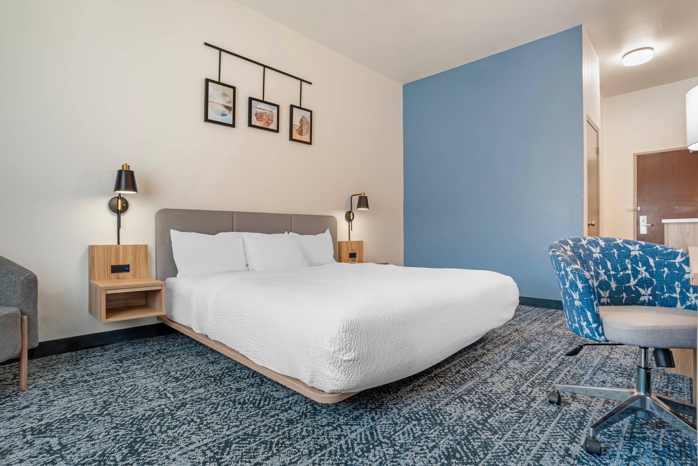Habitació d'hotel amb un llit gran, dues taules de nit amb làmpades, una paret d'accent blau, una catifa estampada i una cadira blava.