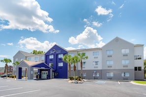 Edificio de Sleep Inn & Suites by Choice Hotels con entrada azul, palmeras y estacionamiento.