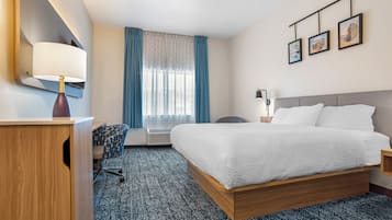 Habitación de hotel con una cama, un escritorio con lámpara, una silla, una ventana con cortinas y una pared adornada con cuadros.
