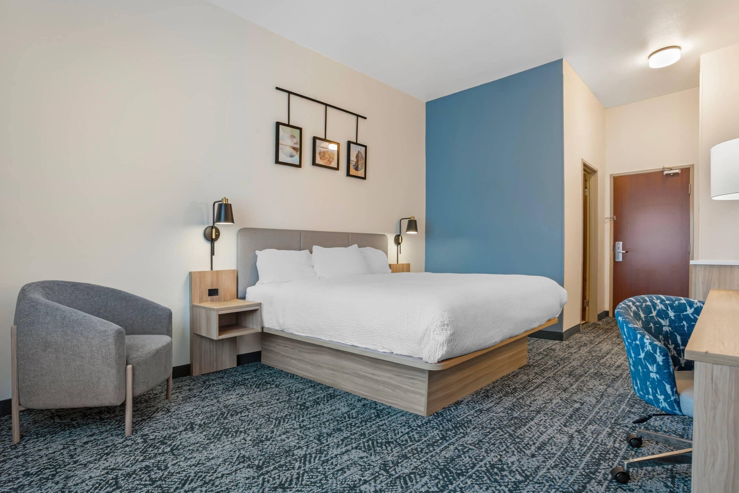 Una habitación de hotel con cama, una silla, un escritorio, una lámpara y un cuadro colgado en la pared.