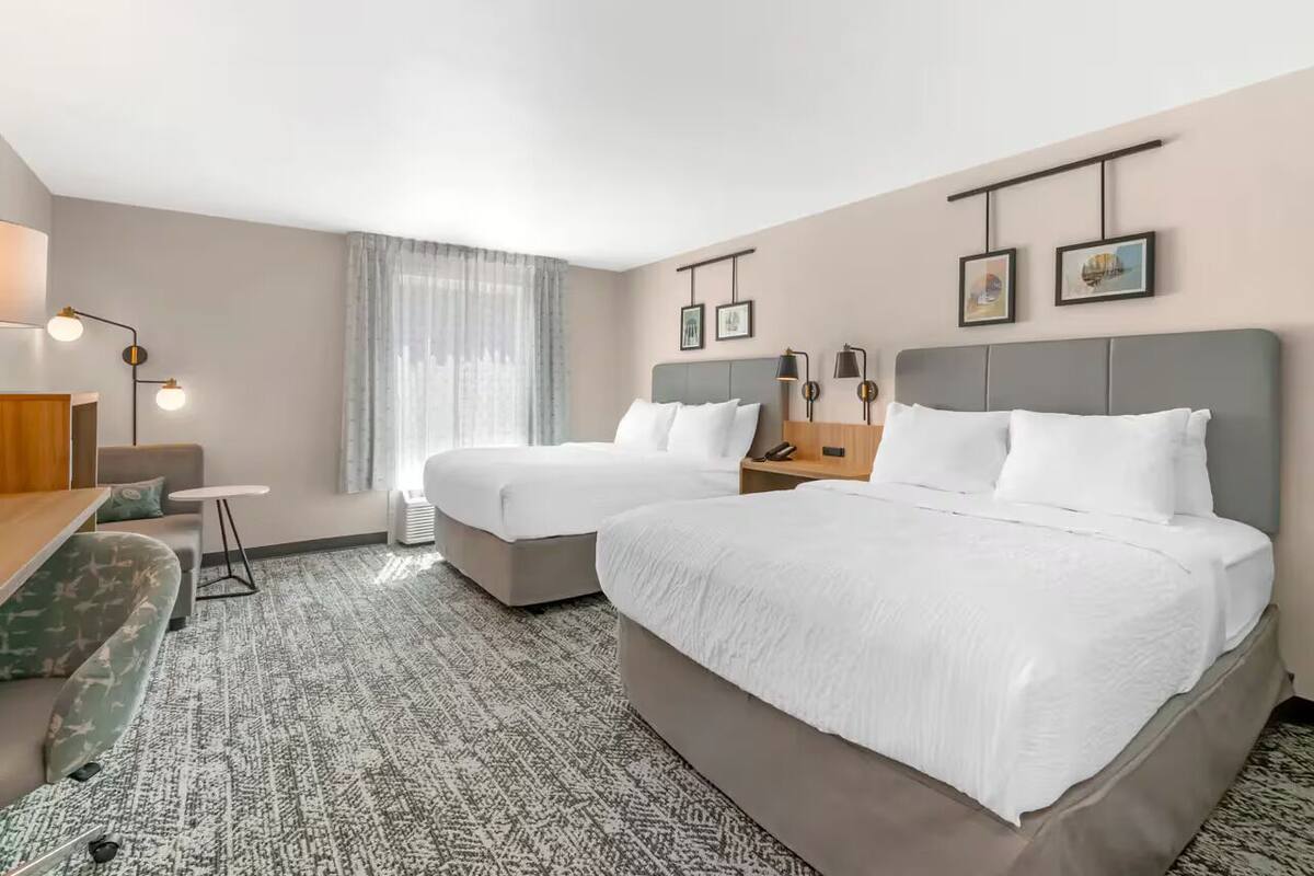 A hotel room with two beds, a sofa, a small table, a lamp, and a window with sheer curtains.