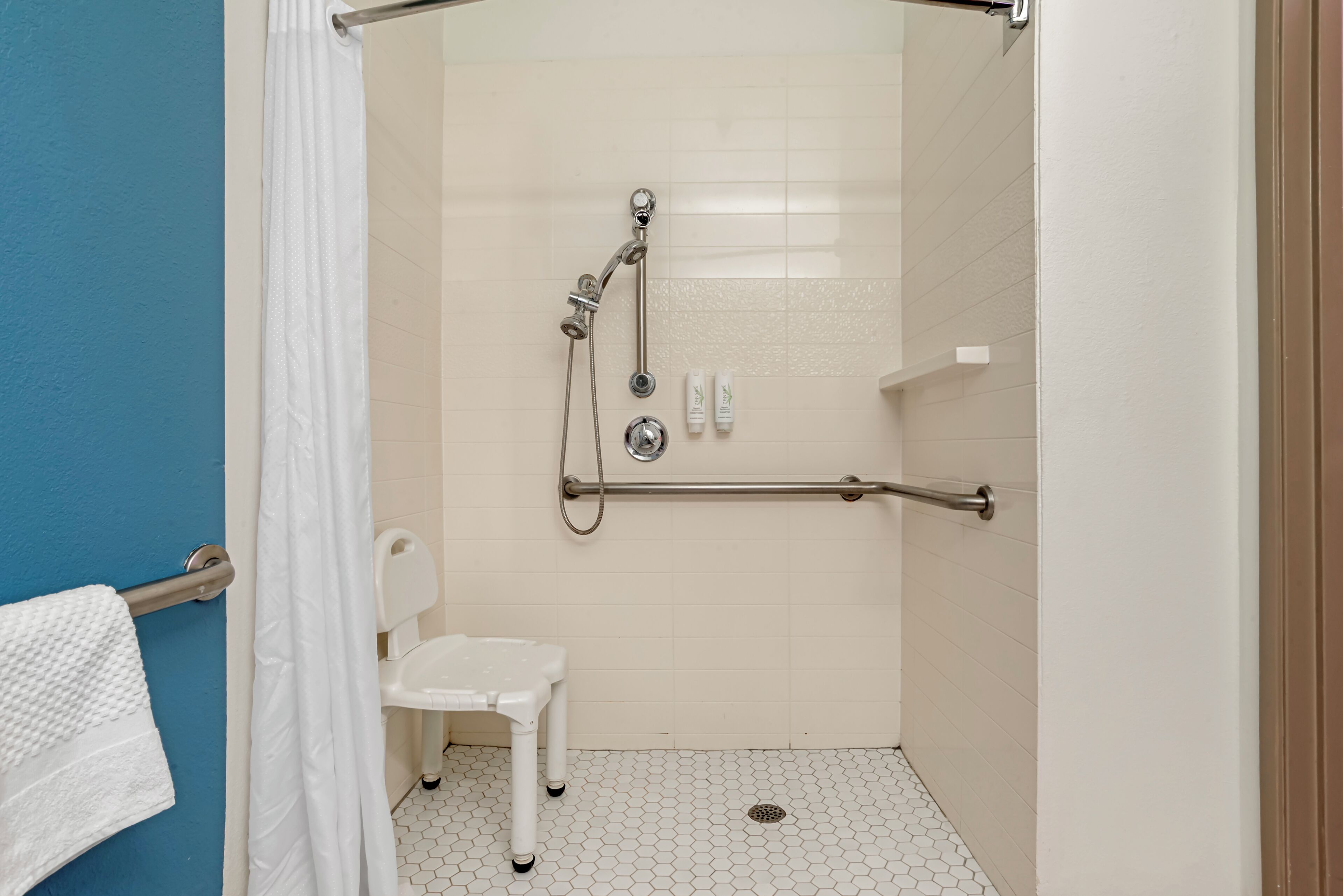 A shower with a grab bar, a white chair, and a toilet.