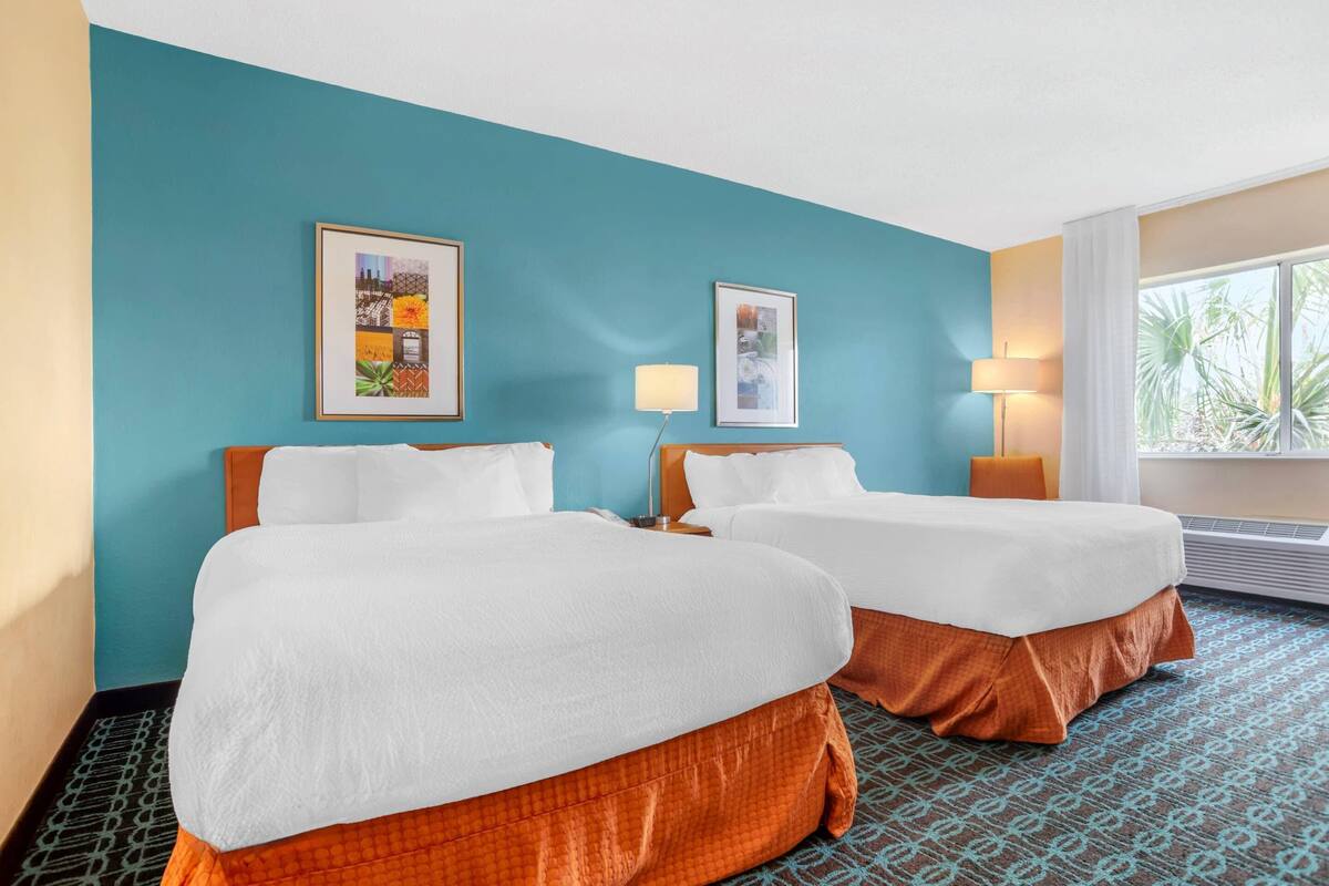A hotel room with two beds, teal walls, framed pictures, and a window with a view.