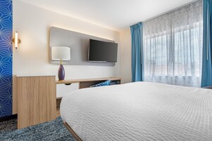 Habitación de hotel con cama, televisor de pantalla plana en la pared, mesita de noche con lámpara y ventana con cortinas.