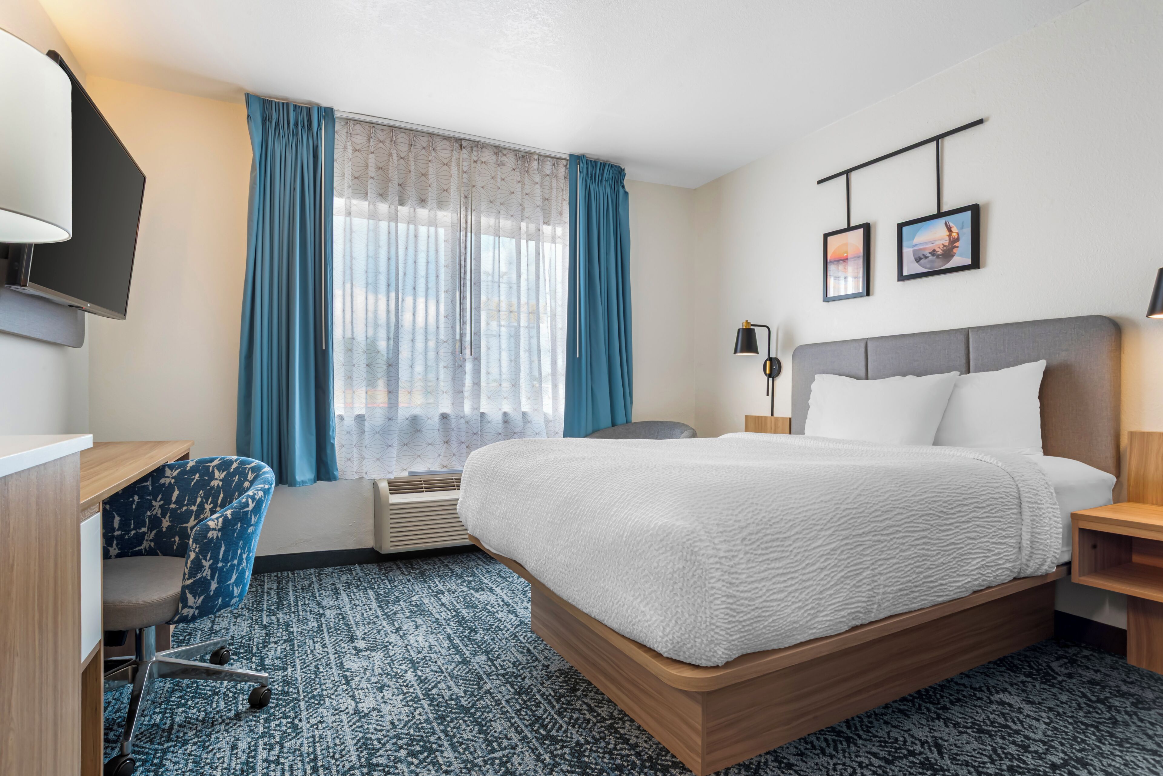 A hotel room with a bed, a desk, a chair, a TV, a window with curtains, and two wall-mounted pictures.