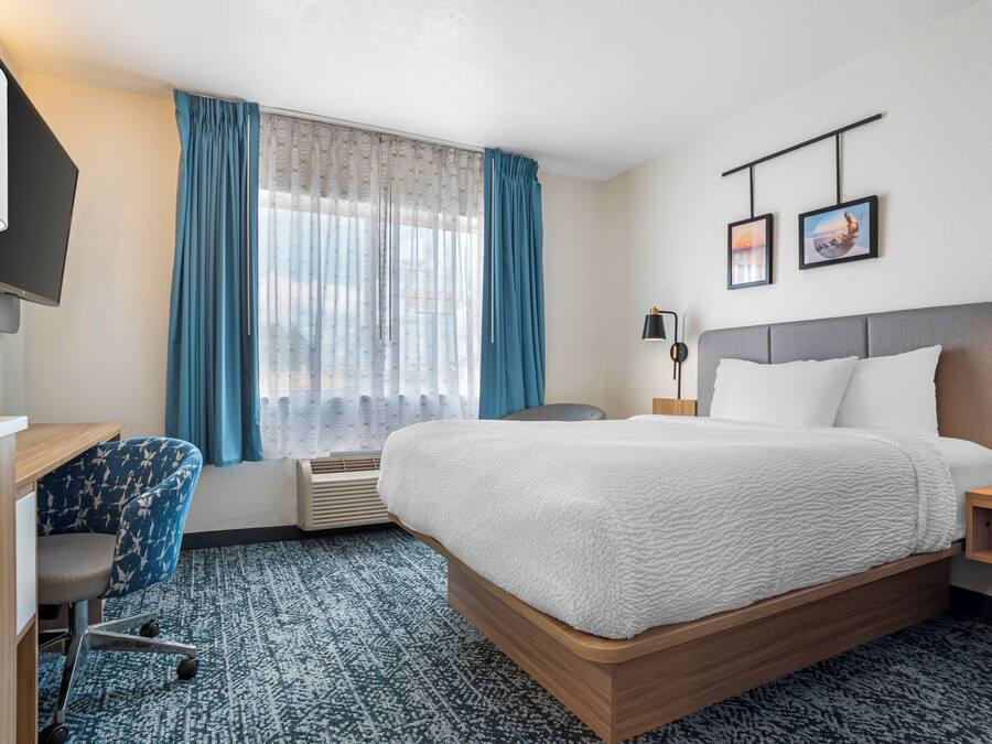 Habitación de hotel con cama, escritorio, silla, televisor, ventana con cortinas y dos cuadros en la pared.