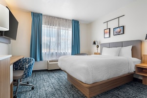 Habitación de hotel con cama, escritorio, silla, televisor, ventana con cortinas y dos cuadros en la pared.