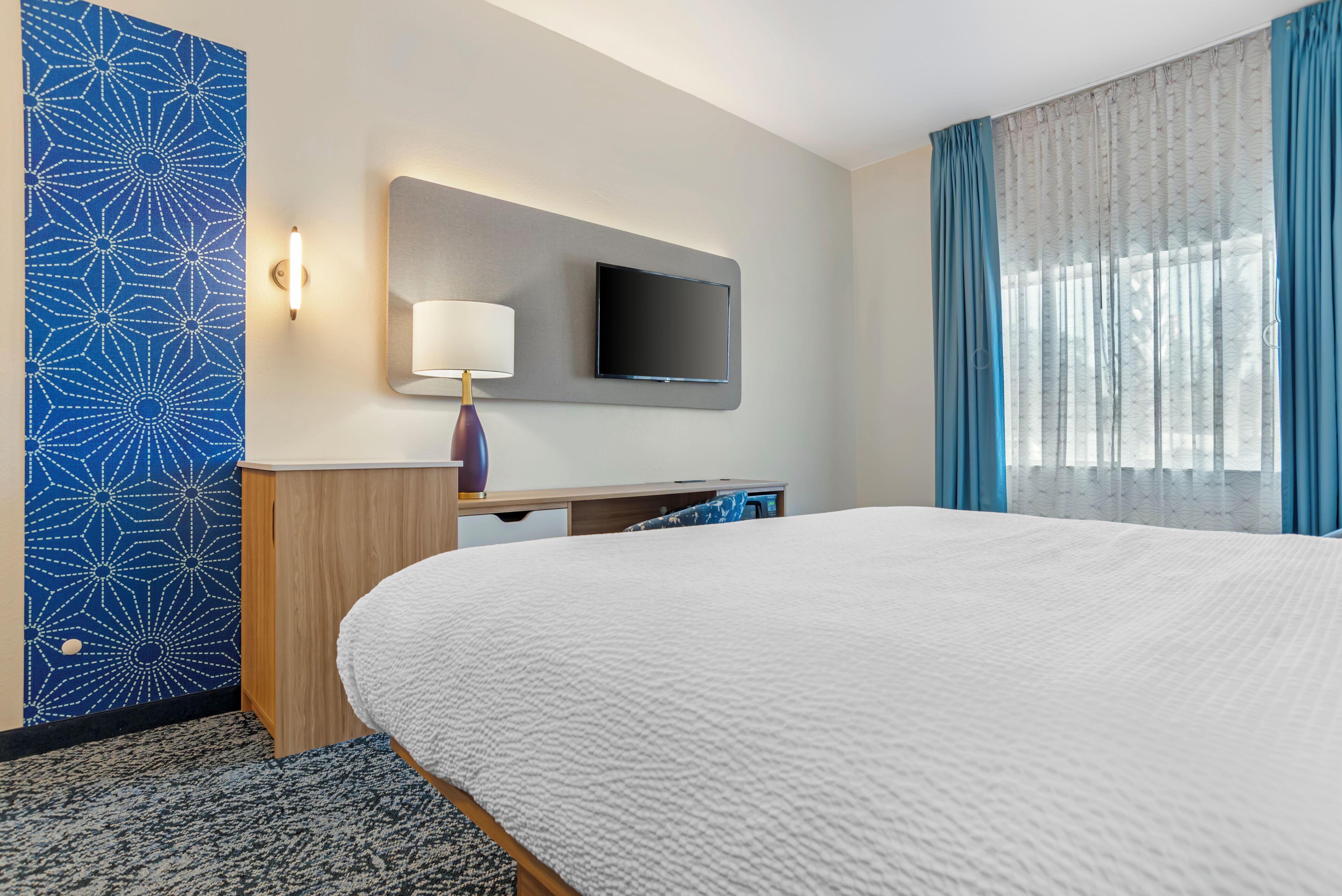 A hotel room with a large bed, a flat-screen TV mounted on the wall, a desk with a lamp, and a window with blue curtains.