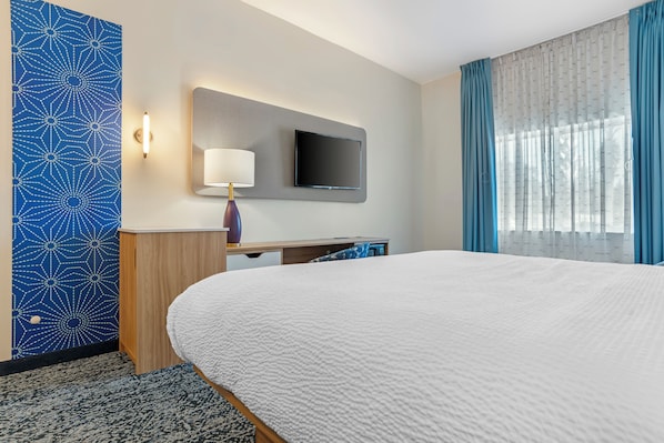 A hotel room with a large bed, a flat-screen TV mounted on the wall, a desk with a lamp, and a window with blue curtains.