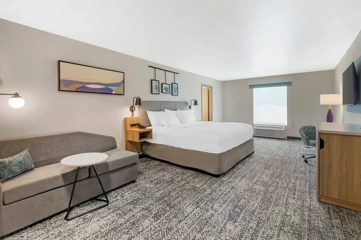 A hotel room with a bed, a sofa, a small table, a desk, a chair, a TV, and a window.