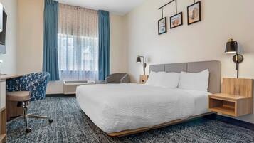 Quarto de hotel com uma cama grande, uma escrivaninha, uma cadeira, abajur e televisão fixada na parede.