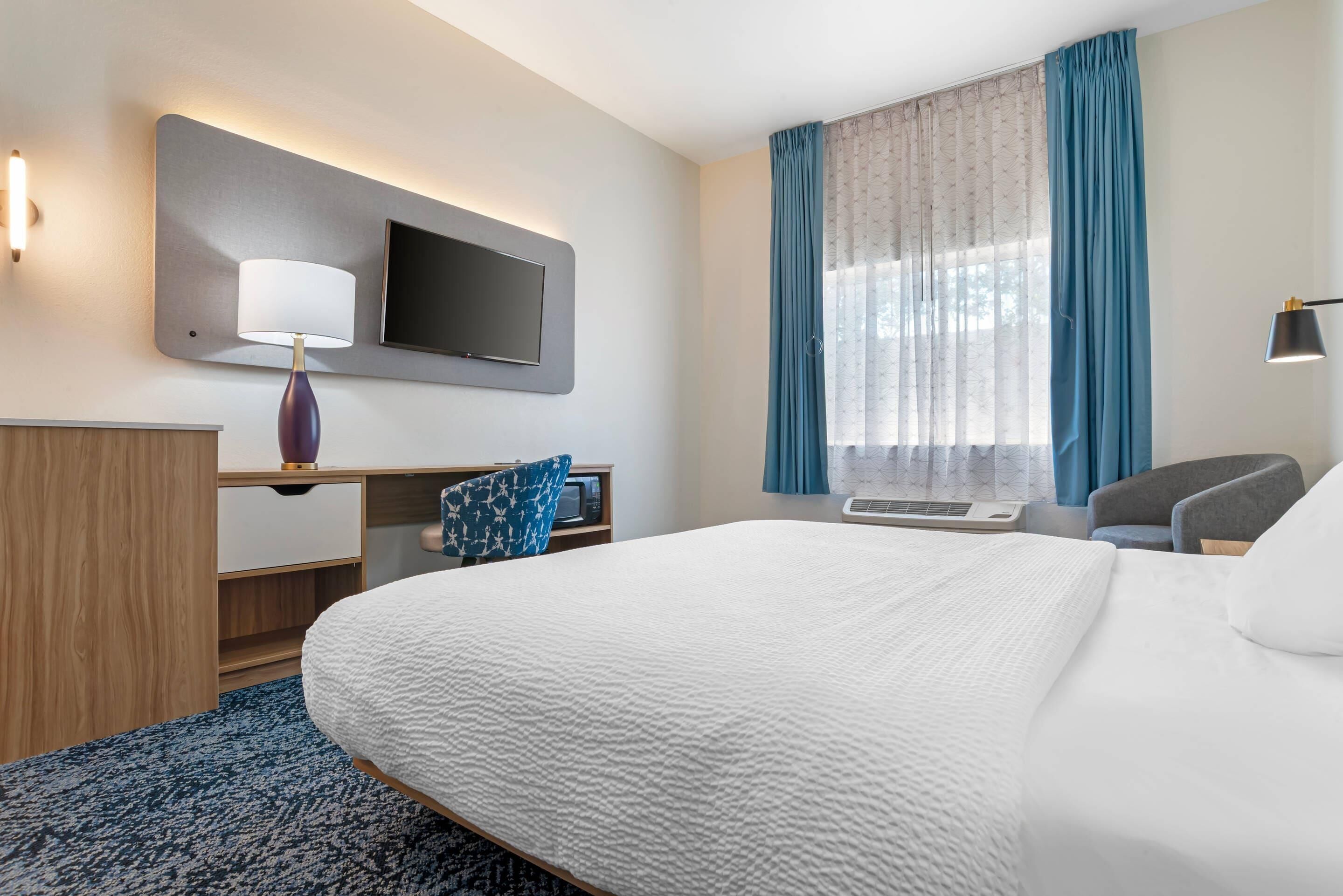 A hotel room with a bed, a desk with a flat-screen TV, a lamp, a chair, and a window with curtains.