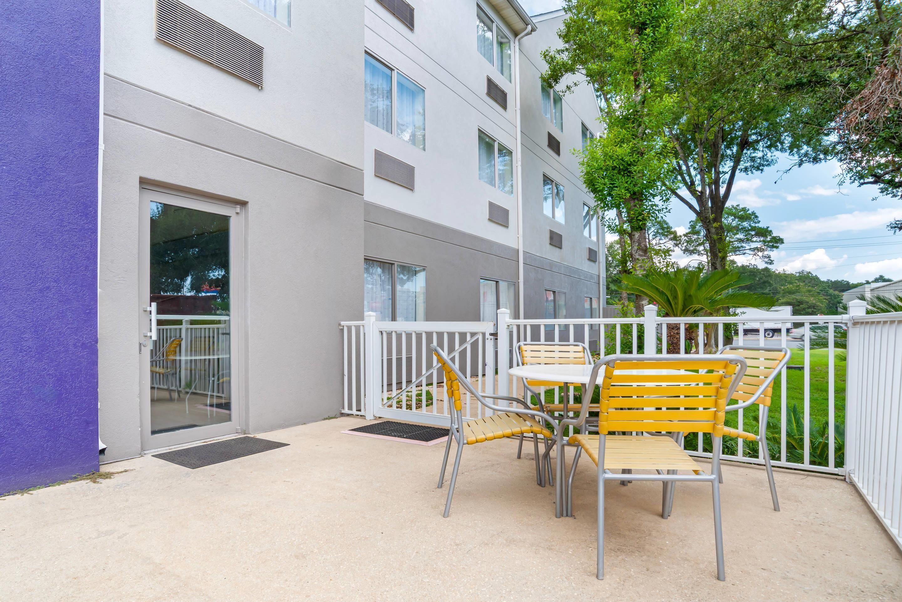 A balcony with a table and chairs, a white railing, and a building with multiple windows and vents.