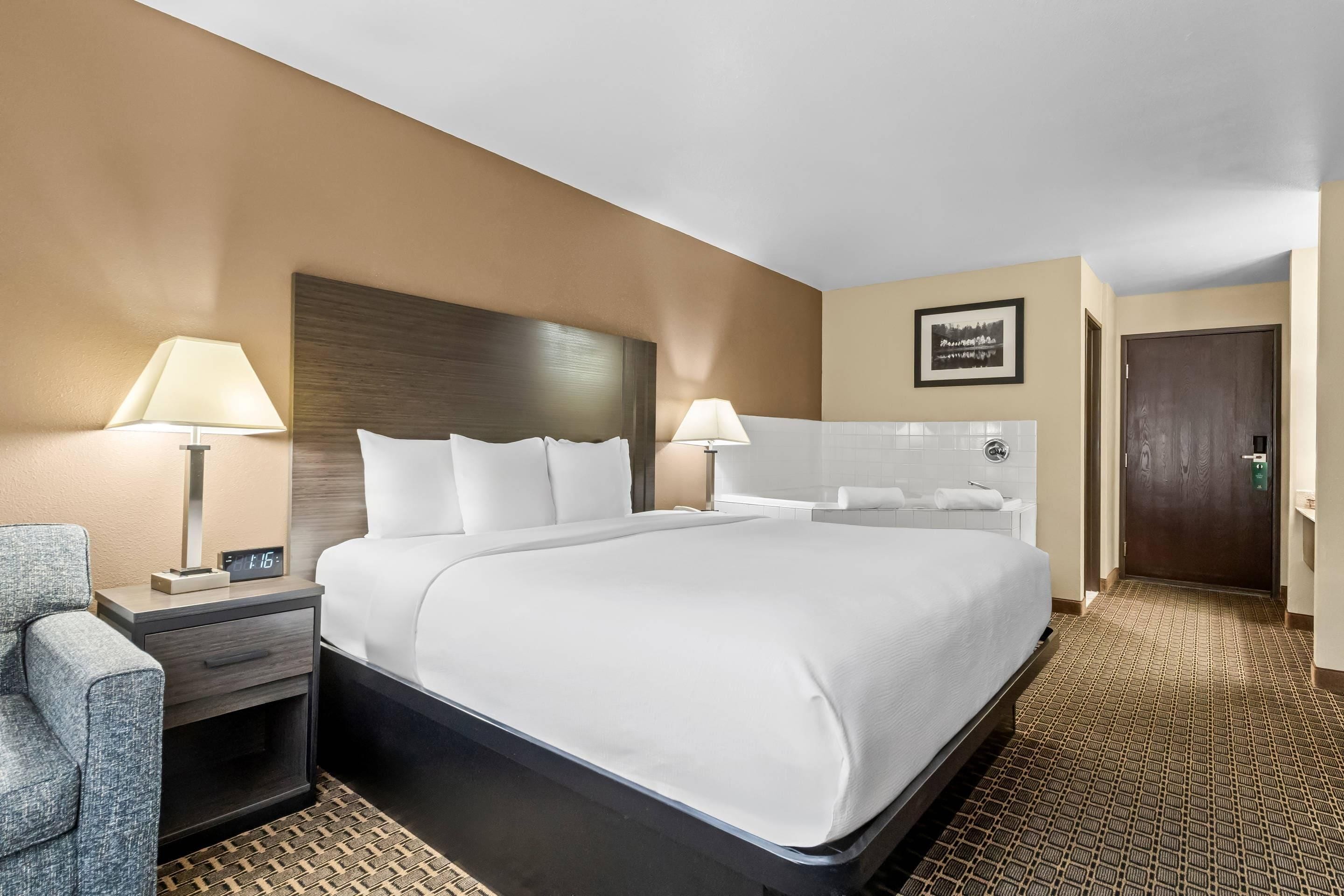 A hotel room with a large bed, bedside lamps, a chair, a nightstand, a framed picture on the wall, and a door.