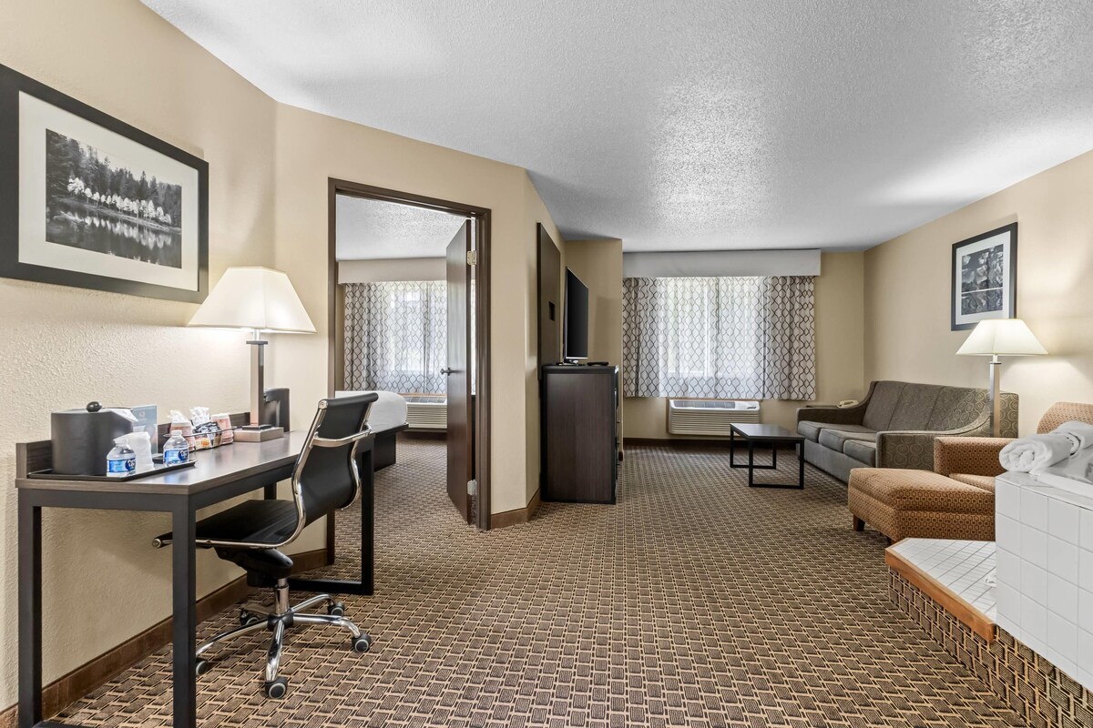 A hotel room with a desk, chair, sofa, and a bed. There are two lamps, a television, and a window with curtains.