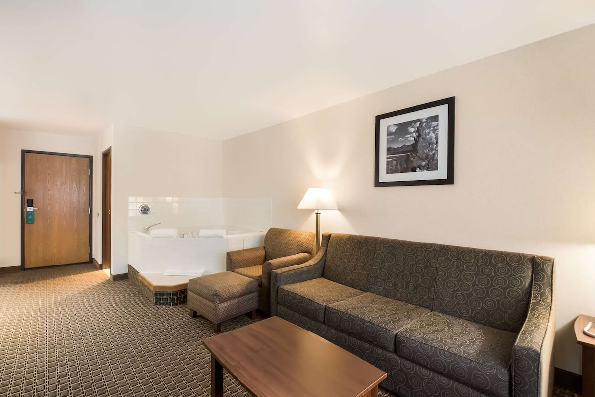 A hotel room with a sofa, armchair, ottoman, and a small table. There is a bathtub and a framed picture on the wall.