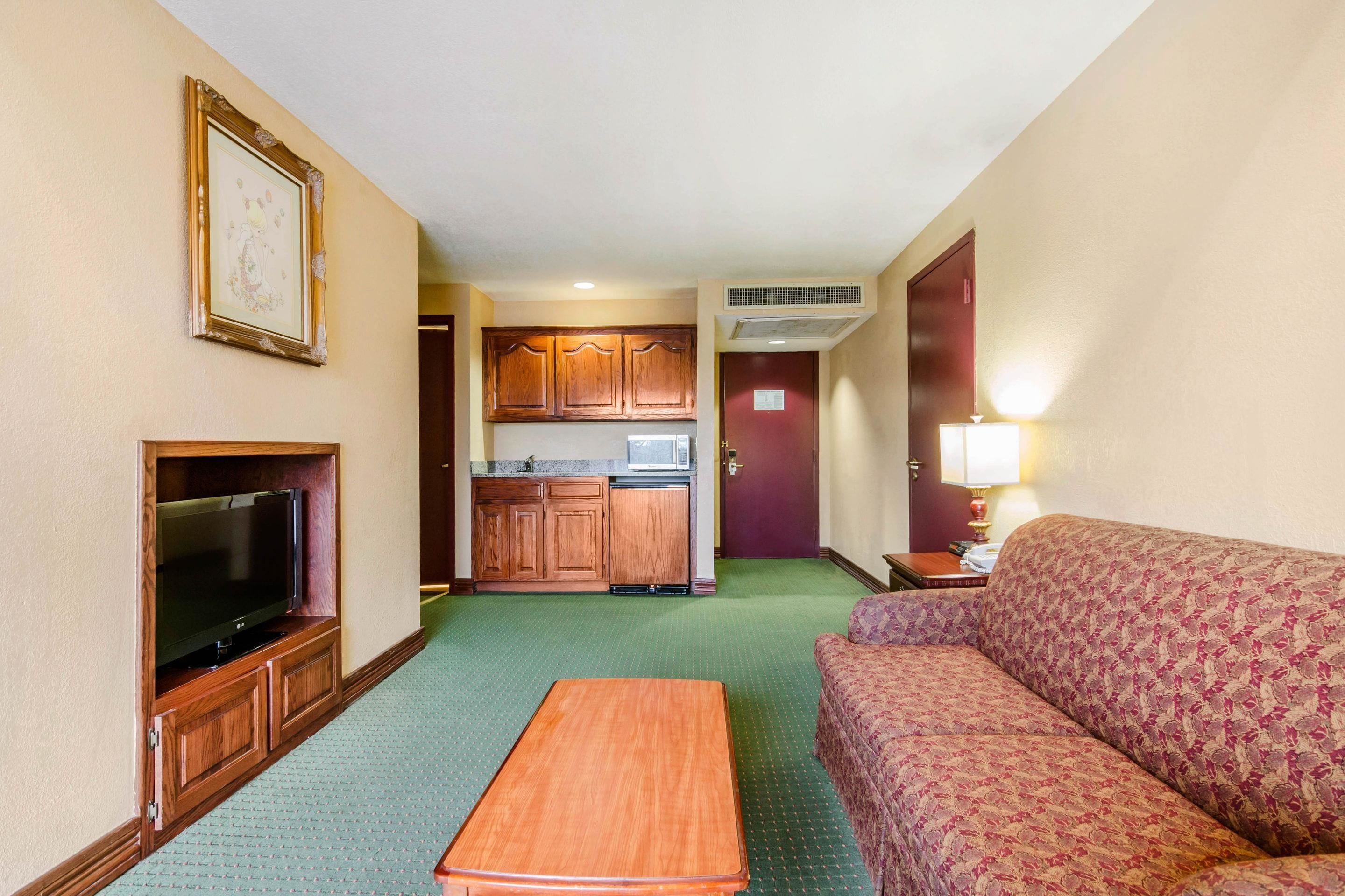 A compact hotel room with a television, a sofa, a small table, a kitchenette, and a framed picture on the wall.