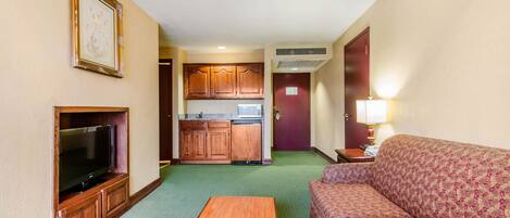 A compact hotel room with a television, a sofa, a small table, a kitchenette, and a framed picture on the wall.