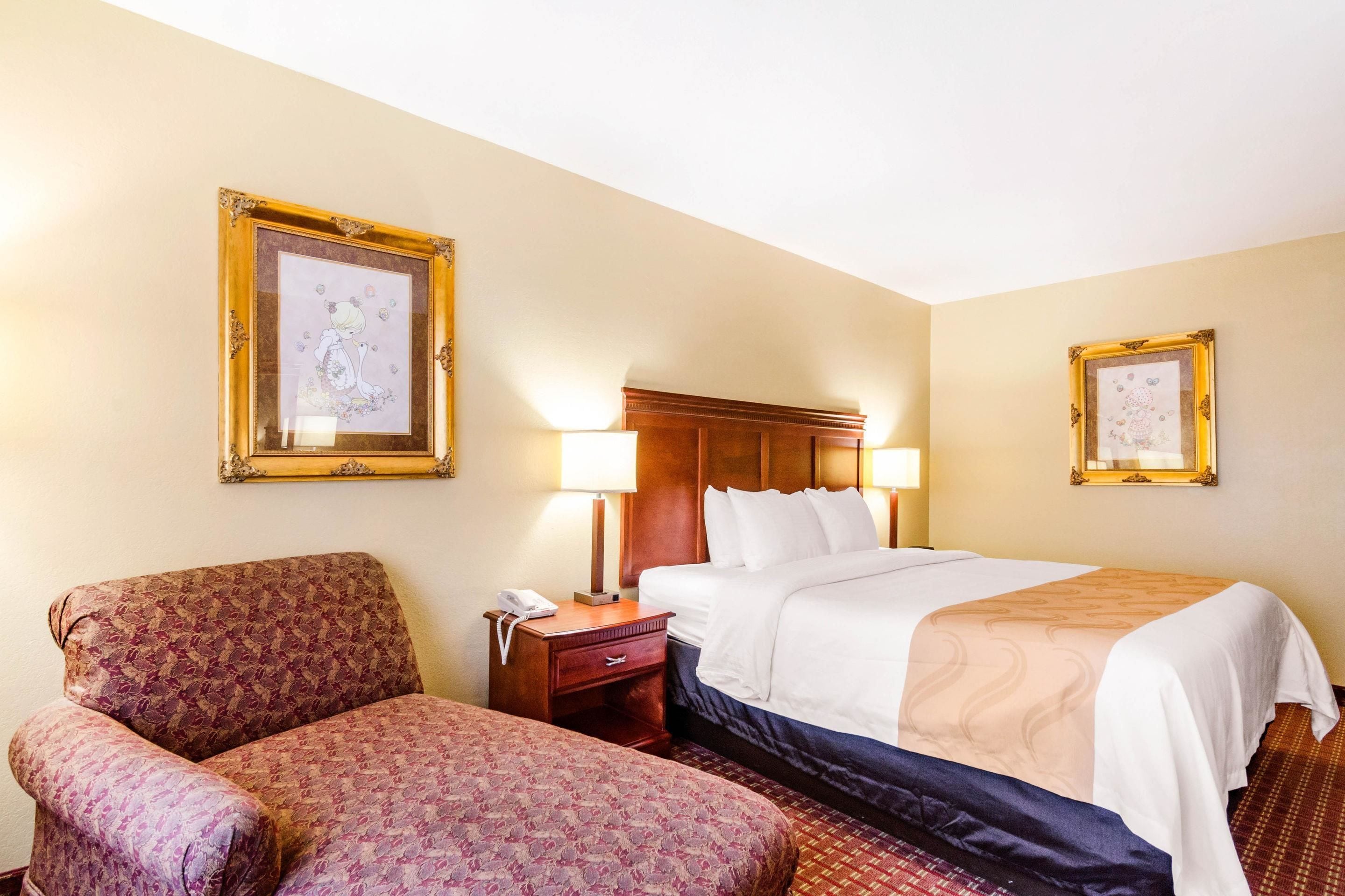 A hotel room with a large bed, a chair, a nightstand with a lamp, and two framed pictures on the wall.