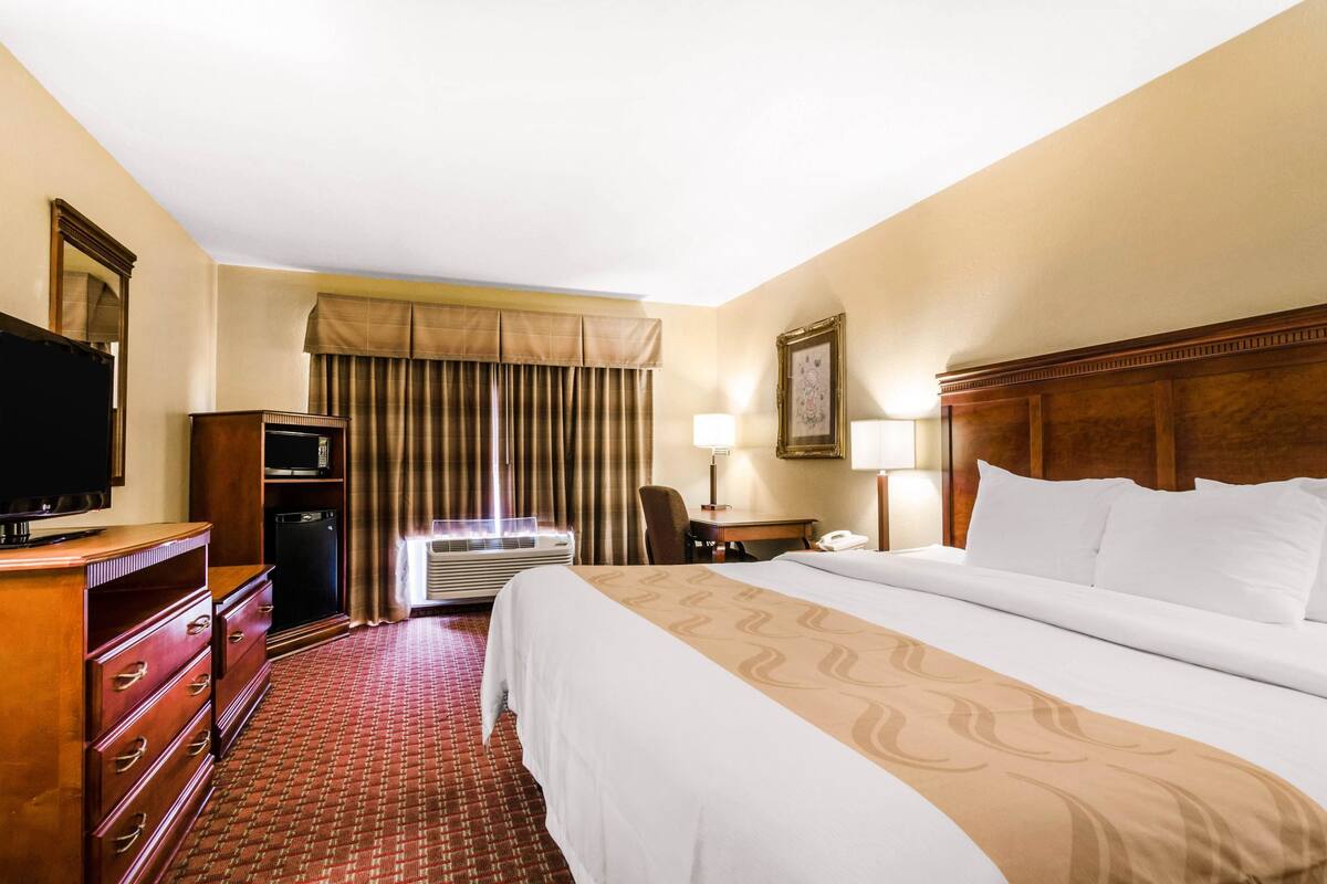 Habitación de hotel con una cama grande, televisión, escritorio con lámpara, una silla y ventana con cortinas.