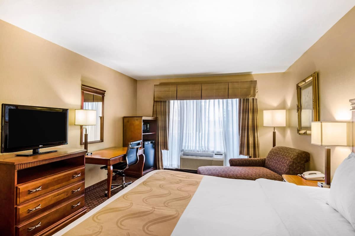 Habitación de hotel con cama, escritorio con computadora, televisión, silla y ventana con cortinas.