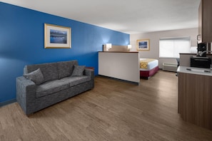 A room with a grey sofa, a bed with a red and yellow bedspread, a kitchenette, and a blue wall with a framed picture.