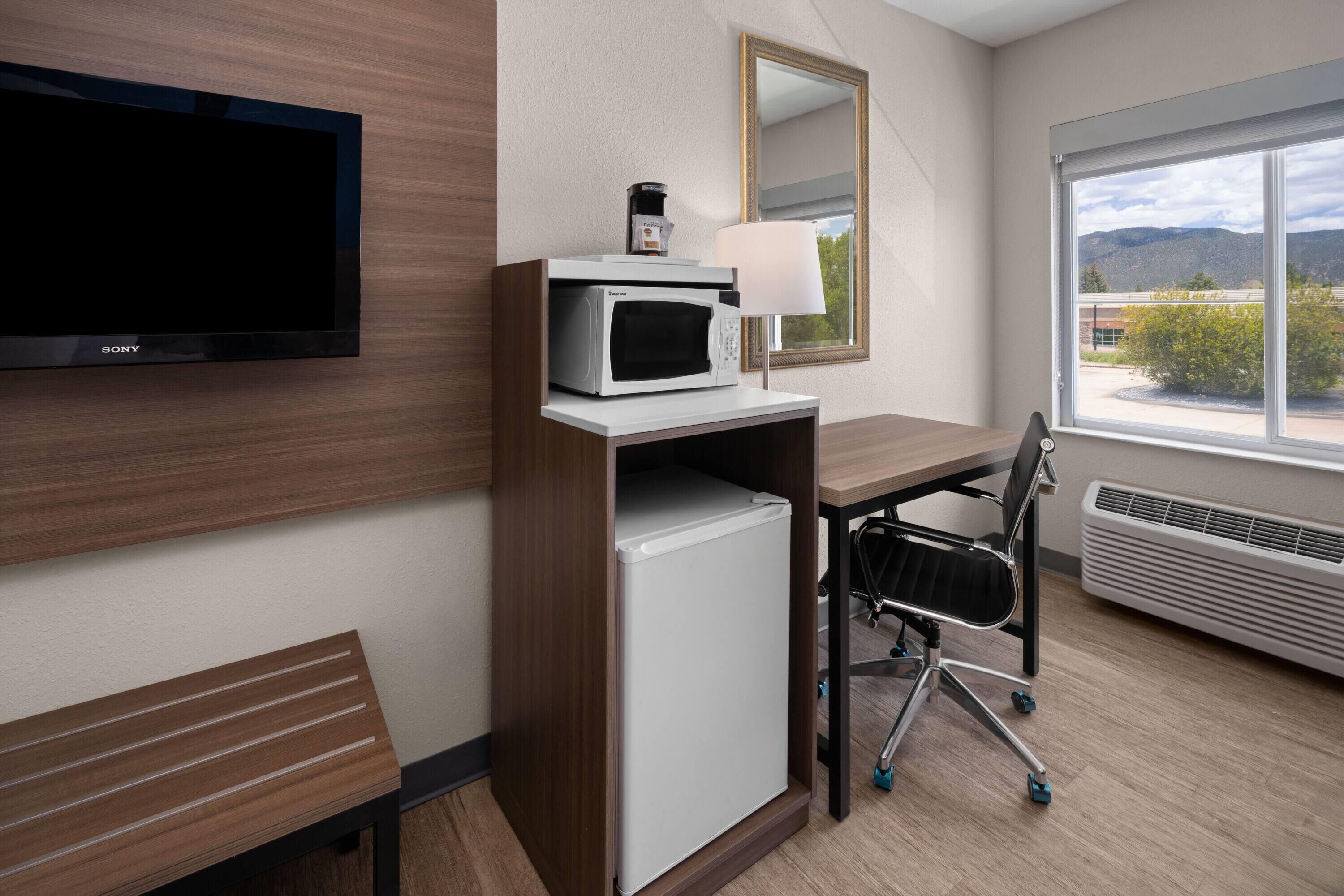 A hotel room with a flat-screen TV, a microwave, a small refrigerator, a desk with a chair, a large mirror, and a window with a view of mountains.