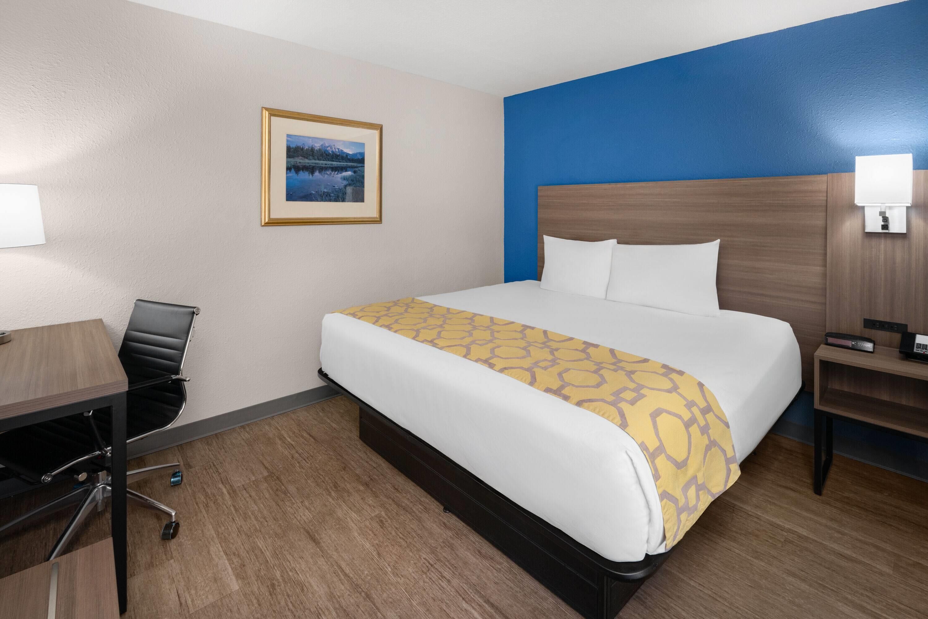 Una habitación de hotel con cama, escritorio, silla, mesita de noche y lámpara de pared.