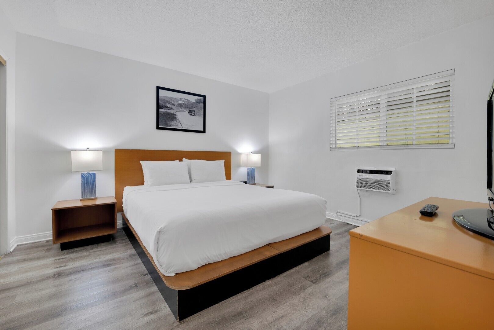 A hotel room with a bed, bedside tables, a desk, a picture on the wall, and a window with blinds.