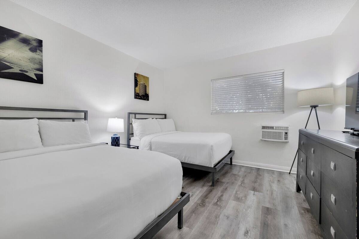 A hotel room with two beds, a dresser, a lamp, a painting, and a window with blinds.