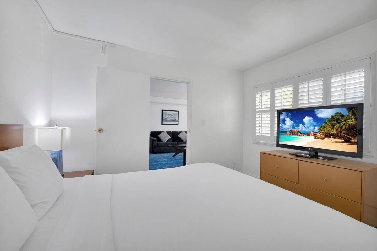A hotel room with a bed, a TV, a dresser, and a door leading to a living area.