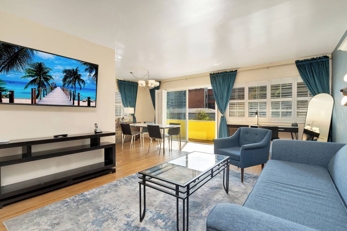 A modern living room with a flat-screen TV, a glass coffee table, a sofa, and a dining area with chairs and a table.