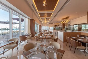 A modern dining area with tables set for service, large windows offering views of the outdoor area, and a kitchen visible in the background.