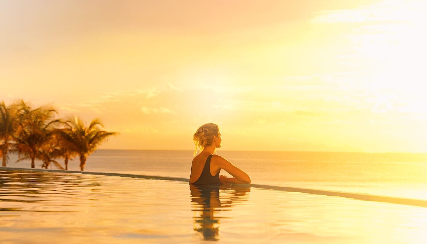 A person in a swimming pool at sunset with palm trees and the ocean in the background.