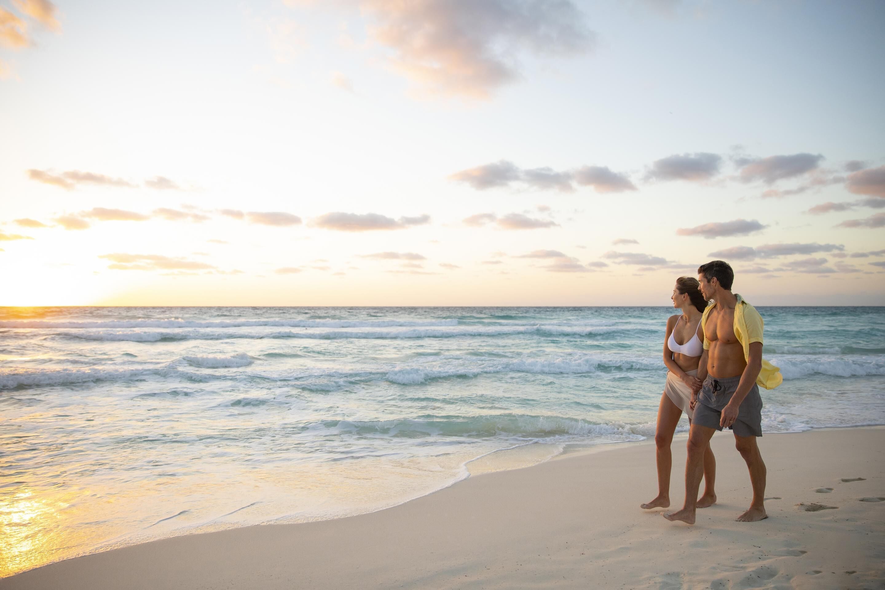 A couple standing on a beach with the ocean and a sunset in the background.