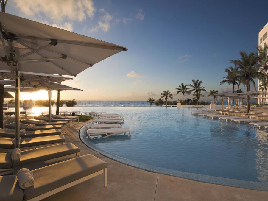A poolside area with sun loungers and umbrellas, a clear view of the ocean, and palm trees.